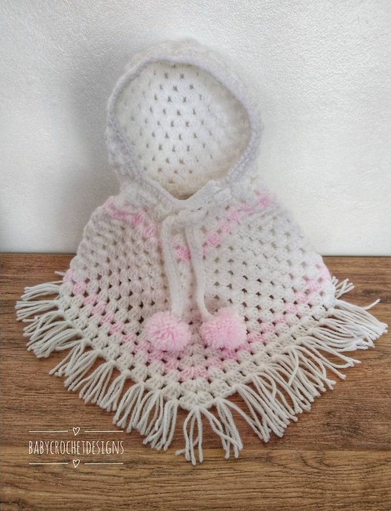 K&ouml;nnte beinhalten: Wei&szlig;es geh&auml;keltes Kapuzenponcho mit rosa Streifen und wei&szlig;en Fransen. Das Poncho hat zwei rosa Bommeln, die an der Kapuze befestigt sind.