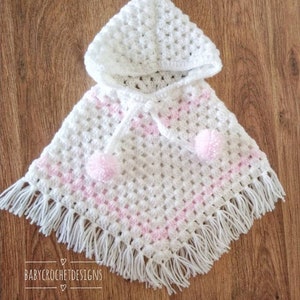 K&ouml;nnte beinhalten: Wei&szlig;es geh&auml;keltes Babyponcho mit rosa Besatz und wei&szlig;en Bommeln. Das Poncho hat eine Kapuze und Fransen.