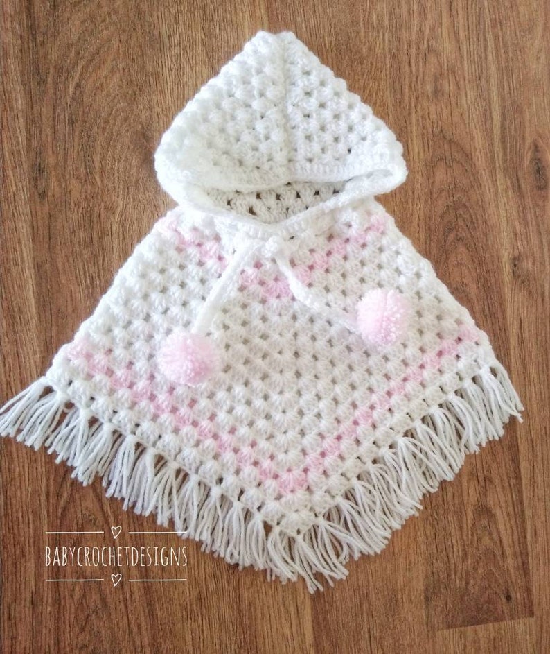 K&ouml;nnte beinhalten: Wei&szlig;es geh&auml;keltes Babyponcho mit rosa Besatz und wei&szlig;en Bommeln. Das Poncho hat eine Kapuze und Fransen.