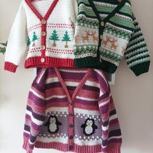 May include: Three handmade crochet cardigans in white, green, and purple. The white cardigan has red trim and Christmas tree designs. The green cardigan has reindeer patterns. The purple cardigan has penguin designs and horizontal stripes. The website address www.babycrochetdesigns.com is visible.