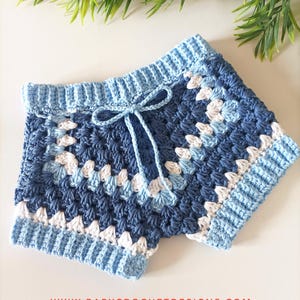 May include: Handmade crochet shorts in shades of blue and white. The shorts feature a drawstring waist and a textured stitch pattern. The design includes a light blue waistband and cuffs, with a repeating pattern of white and darker blue squares.