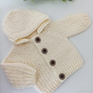 May include: A cream-colored crocheted baby cardigan with a hood and wooden buttons. The cardigan is made with a textured stitch and has a simple design.