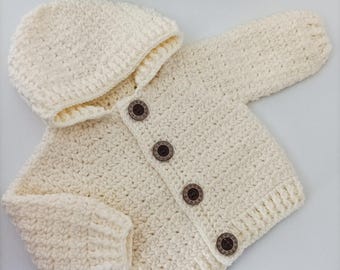Könnte beinhalten: Ein cremefarbener gehäkelter Baby-Cardigan mit Kapuze und Holz-Knöpfen. Der Cardigan ist mit einem strukturierten Stich gearbeitet und hat ein schlichtes Design.