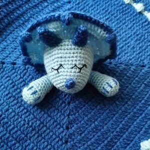 May include: A blue crocheted baby blanket with a light blue crocheted dinosaur attached. The dinosaur has closed eyes and a small blue nose.
