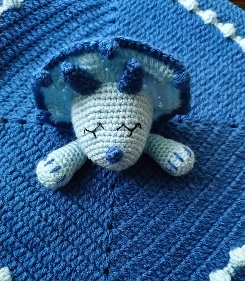 May include: A blue crocheted baby blanket with a light blue crocheted dinosaur attached. The dinosaur has closed eyes and a small blue nose.