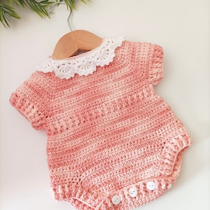 Op de afbeelding: Een gehaakte baby romper in een perzikroze kleur met een witte gehaakte kraag. De romper heeft korte mouwen en een drukknoopsluiting aan de onderkant. De romper hangt aan een houten hanger.