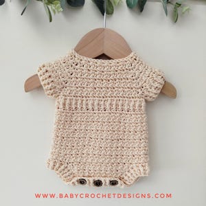 May include: A crocheted baby onesie in a light beige color with short sleeves and three brown buttons. The onesie is hanging on a wooden hanger against a white background.