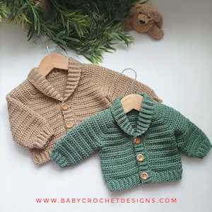 May include: Two crocheted baby cardigans, one in a light brown color and one in a dark green color, with wooden buttons and a collared neckline. The cardigans are hanging on wooden hangers.
