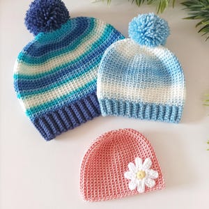 May include: Three crocheted hats, one blue and white striped with a blue pompom, one light blue and white ombre with a light blue pompom, and one pink with a white daisy.