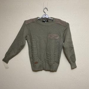 Może przedstawiać: Szałwiowy zielony sweter z teksturowanym wzorem dzianiny, zamszowymi naszywkami na ramionach i kieszenią na piersi. Sweter z długim rękawem wisi na metalowym wieszaku. Marka "MiCho" jest widoczna na metce.