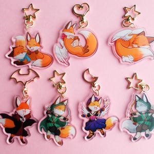 Puede incluir: Conjunto de ocho abalorios acrílicos de colores con zorros en varias poses. Cada abalorio tiene un anillo de metal dorado y una parte superior en forma de estrella. Los zorros están representados en diferentes colores, incluyendo naranja, blanco y gris.