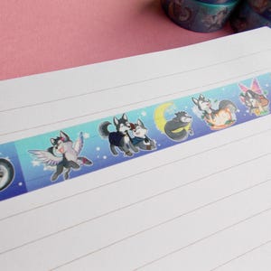 Op de afbeelding: Een blauwe en witte washi tape met een herhalend patroon van cartoon husky honden in verschillende poses. De honden dragen verschillende accessoires, zoals vleugels, hoeden en muziekinstrumenten. De tape is op een wit notitieboek met gestreept papier.