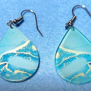 May include: A pair of teardrop-shaped earrings featuring a translucent light blue base with swirling white and darker blue marbled patterns. Each earring is attached to a silver-toned metal fishhook ear wire for hanging.