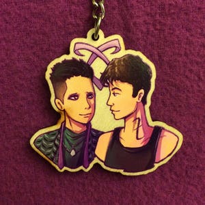 May include: A wooden keychain featuring two men with brown hair. The man on the left is wearing a green and black jacket with a silver necklace. The man on the right is wearing a black tank top. The keychain has a purple ribbon design at the top.