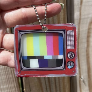 May include: A square-shaped acrylic keyring featuring a red vintage television set with a screen displaying a colourful vertical test pattern and a horizontal greyscale bar. The television includes two circular knobs and a speaker grille on the right side.