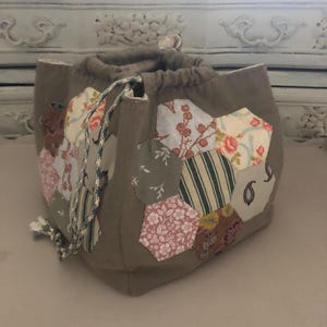 Könnte beinhalten: Ein grauer Beutel mit Kordelzug und Patchwork-Design. Der Patchwork-Stil zeigt verschiedene Blumen- und Streifenmuster in Rosa, Grün und Creme. Die Tasche hat einen Kordelzugverschluss mit einer gestreiften Kordel.