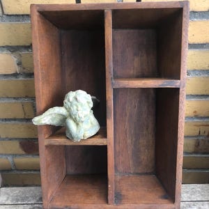 Könnte beinhalten: Eine dunkle Holzaufbewahrungsbox mit vier Fächern und einem schwarzen Metallgriff. Eine kleine, hellfarbene Engelsstatue ruht auf einem der Regale. Die Box ist aus dunkelbraunem Holz gefertigt.