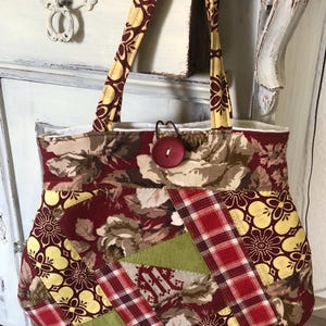 Könnte beinhalten: Eine Patchwork-Tragetasche mit rotem, grünem und gelbem Blumendruck. Die Tasche hat einen roten Knopf und zwei braune Henkel.
