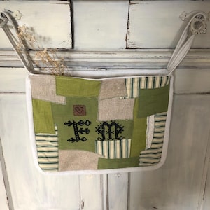 Könnte beinhalten: Ein grüner und weißer Patchwork-Stoff-Hängeregal mit weißem Besatz und zwei weißen Schlaufen. Der Organizer hat das Wort "Home" in schwarzem Faden auf der Vorderseite gestickt.