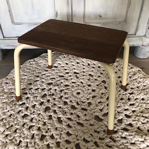 Könnte beinhalten: Ein kleiner, vintage Tritthocker mit einer rechteckigen, dunkelbraunen Holzfläche. Die Beine sind cremefarbenes Metall mit braunen Gummifüßen. Der Hocker steht auf einem cremefarbenen, gehäkelten Teppich.