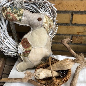 Könnte beinhalten: Eine handgefertigte Stoff-Seepferdchen-Puppe mit Patchwork-Design, floralen Akzenten und einem braunen Knopfauge. Ein kleiner Weidenkorb enthält zwei Stoffkarotten. Ein weißer Kranz und Holzäste vervollständigen die rustikale Szene.