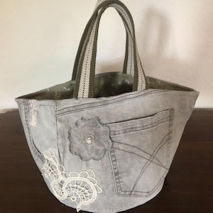 Könnte beinhalten: Graue Jeans-Tote-Tasche mit floraler Applikation und Spitzendetails. Die Tasche hat eine Tasche, olivgrüne und cremefarbene gestreifte Griffe und ein floral gemustertes Innenfutter. Die Tasche ist handgefertigt.
