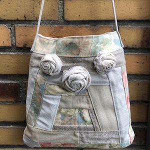 Könnte beinhalten: Eine Patchwork-Schultertasche aus Stoff mit hellgrauem Hintergrund und Blumenmustern. Die Tasche hat drei große Stoffrosetten in hellgrauer Farbe.