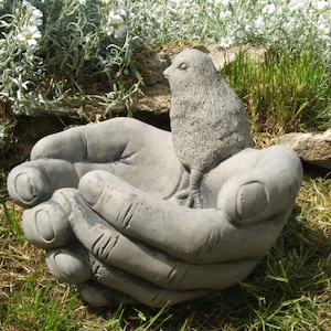 May include: A concrete bird bath shaped like two hands cupping a small bird. The bird bath is gray and has a rough texture.