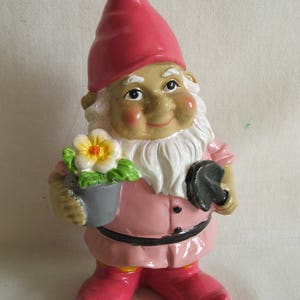 May include: A pink garden gnome figurine with a white beard, holding a grey pot with a white flower and a black shovel. The gnome is wearing a pink hat, a pink shirt, a black belt, and pink boots.