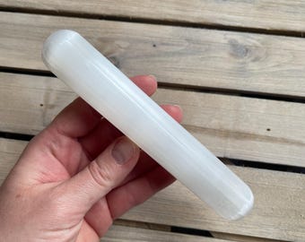 Puede incluir: Una varita de selenita blanca, de forma cilíndrica, sostenida en una mano. La varita es lisa y opaca, con una punta ligeramente redondeada. El fondo es una superficie de madera.