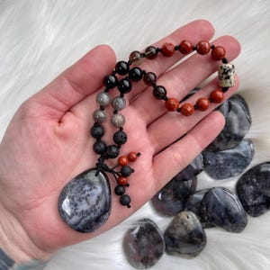Puede incluir: Un collar con un colgante de ágata negro y gris, jaspe rojo, obsidiana negra, cuarzo ahumado, roca volcánica y un cordón negro. El collar se muestra en una mano con varias piedras de ágata sueltas.