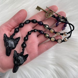 Puede incluir: Pendientes de cabeza de lobo de obsidiana negra en un cordón negro con cuentas de amatista, estampado de leopardo y cuarzo transparente. Un dije de llave dorada cuelga del cordón.