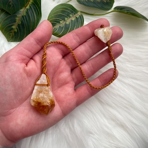 Pode incluir: Um pingente de cristal de citrino com um cordão trançado marrom. O cristal é de cor amarelo-alaranjado claro, e o cordão é amarrado ao redor do cristal. O pingente é segurado em uma mão.