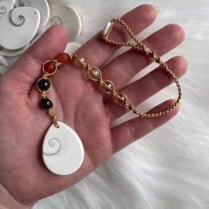 May include: A white shell pendant with a spiral pattern hangs from a brown cord with beads in shades of red, brown, and clear.