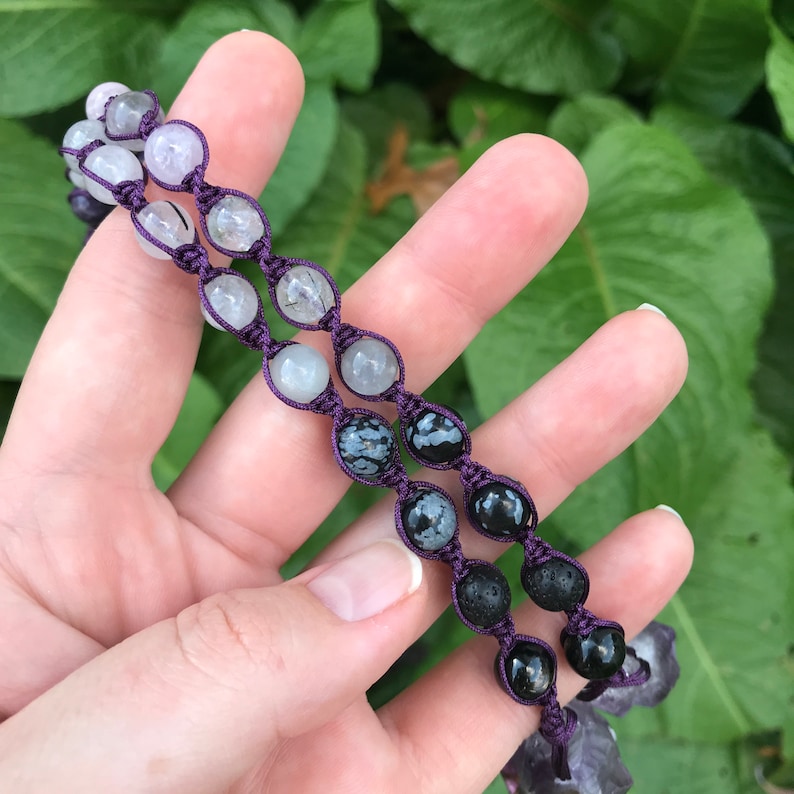 Puede incluir: Dos pulseras de cuentas con un cord&oacute;n morado. Las cuentas son una mezcla de piedras transparentes, blancas, negras y grises moteadas. Las pulseras se sostienen en una mano sobre un fondo de hojas verdes.