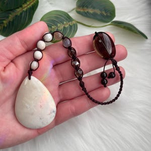May include: A handcrafted pendant necklace featuring a large, teardrop-shaped white stone. The necklace includes a series of white and brown beads, and a dark brown, oval-shaped stone. The stones are connected by a dark brown, woven cord.