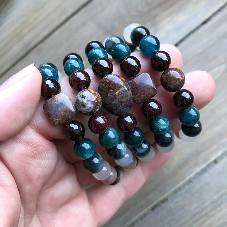 May include: A stack of bracelets with various gemstone beads, including green, blue, brown, and red beads. The bracelets feature a variety of shapes and sizes of beads.