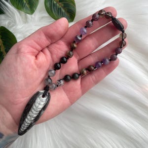 May include: A necklace featuring a dark, elongated ammonite fossil pendant with visible white spiral chambers, suspended from a black cord strung with a variety of round, polished gemstone beads in shades of purple, brown, black, and clear quartz.