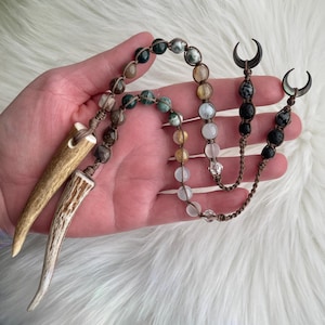 May include: A handcrafted necklace featuring a variety of gemstone beads in shades of green, brown, yellow, and clear. The necklace is accented with two antler pendants and two crescent moon charms, all connected by a brown cord.