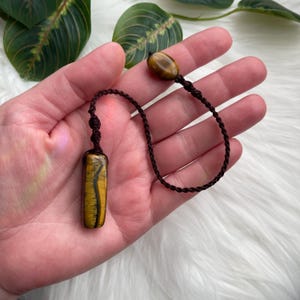 May include: A tiger's eye pendant with a rectangular, yellow and brown stone. The pendant is attached to a brown, braided cord with a matching bead at the end. The cord is knotted at both ends of the pendant.