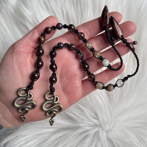 May include: A handmade necklace featuring a series of gemstone beads in shades of red, brown, and cream, strung on black cord. The necklace is adorned with antique brass snake charms and teardrop-shaped pendants.