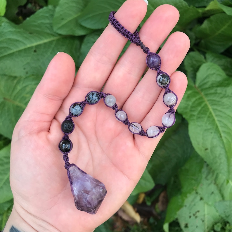 Puede incluir: Un collar de macram&eacute; morado con un colgante de cristal de amatista en bruto. El collar incluye una serie de cuentas de piedras preciosas en varios colores, incluyendo negro, blanco y verde. Las cuentas est&aacute;n ensartadas con un cord&oacute;n de macram&eacute; morado.
