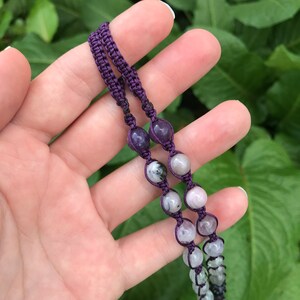 Puede incluir: Primer plano de un collar de macram&eacute; morado hecho a mano con varias cuentas de piedras preciosas. Las cuentas son en tonos morados, grises y blancos. El collar se muestra sobre un fondo de hojas verdes.