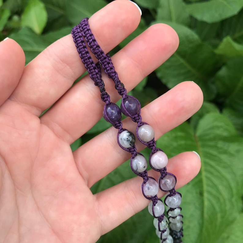 Puede incluir: Primer plano de un collar de macram&eacute; morado hecho a mano con varias cuentas de piedras preciosas. Las cuentas son en tonos morados, grises y blancos. El collar se muestra sobre un fondo de hojas verdes.