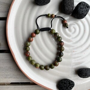 May include: A green and red gemstone bracelet with a black cord closure. The bracelet is on a white plate with black stones.