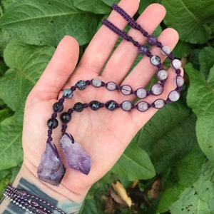 Puede incluir: Un collar de macram&eacute; morado con una variedad de cuentas de piedras preciosas, incluyendo piedras negras, transparentes y moteadas. Dos colgantes de cristal de amatista en bruto cuelgan de la parte inferior. El collar se muestra sobre un fondo de follaje verde.