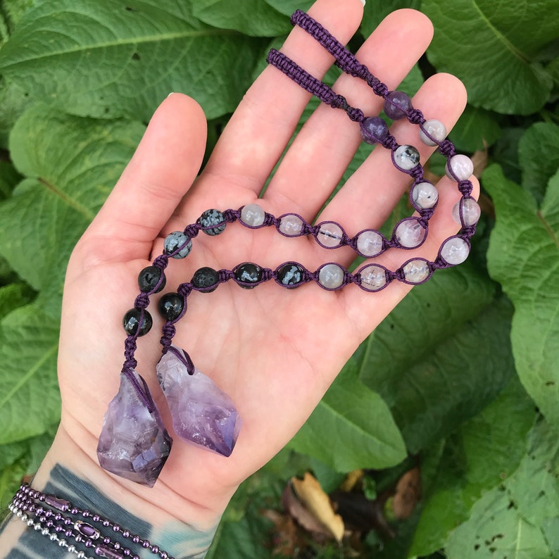 Puede incluir: Un collar de macram&eacute; morado con una variedad de cuentas de piedras preciosas, incluyendo piedras negras, transparentes y moteadas. Dos colgantes de cristal de amatista en bruto cuelgan de la parte inferior. El collar se muestra sobre un fondo de follaje verde.