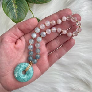 Puede incluir: Un collar mala de piedras preciosas rosa claro, azul y verde con un gran colgante de aventurina verde. El collar está hecho con piedras naturales y está encordado en un cordón delgado.