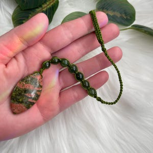 May include: A handmade necklace featuring a teardrop-shaped pendant with red and green patterns. The pendant is connected to a string of green beads and a green woven cord. The necklace is held in a hand.
