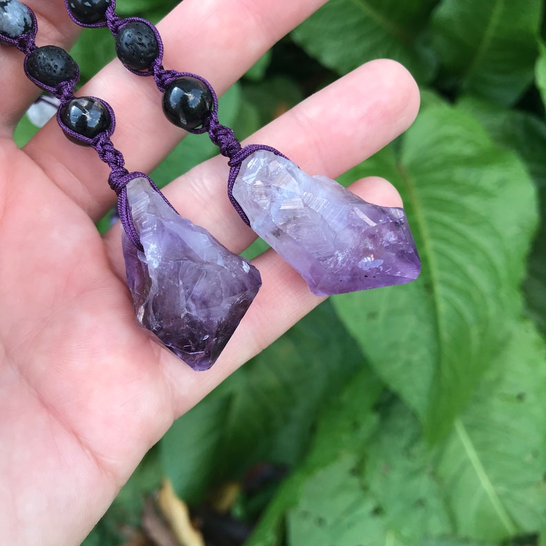 Puede incluir: Dos colgantes de cristal de amatista con un cord&oacute;n morado oscuro y cuentas de lava negras. Los cristales de amatista en bruto presentan un degradado de tonos morados, desde el violeta intenso hasta el lavanda p&aacute;lido. El cord&oacute;n est&aacute; intrincadamente anudado, y las cuentas son lisas y redondas.